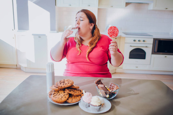 Fat young woman in kitchen sitting and eating sweet food. Biting chocolate. Cookies and cakes on table. Body positive. Daylight. - 写真・画像