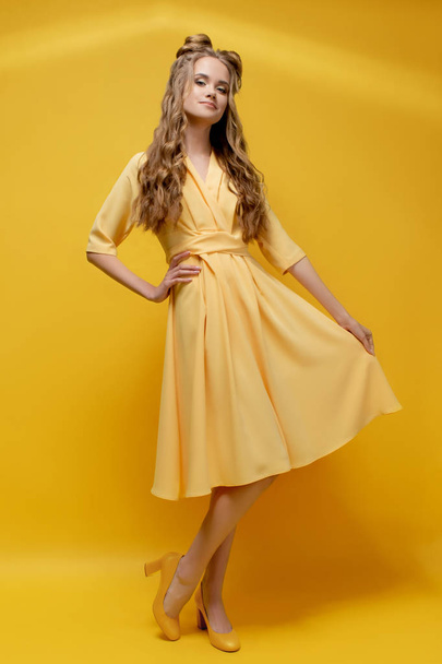A cute young girl in a yellow dress, in shoes with heels on a yellow background with a haircut and curly long hair. Yellow fashionable, stylish, youth clothing. Salon hairstyles and makeup. - Foto, Imagem