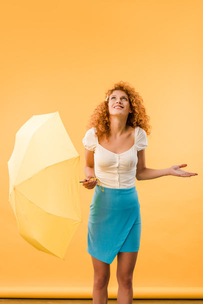 привлекательная рыжая женщина позирует с зонтиком, изолированным на желтом
 - Фото, изображение