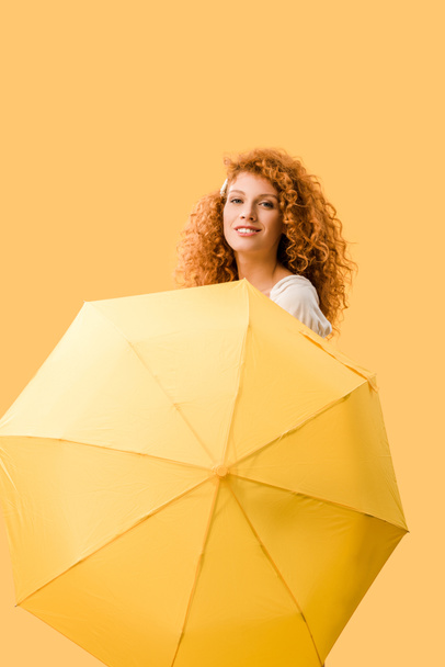 happy redhead woman posing with umbrella isolated on yellow  - Fotó, kép