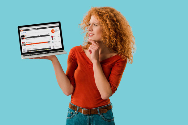 KYIV, UKRAINE - JULY 16, 2019: pensive redhead woman holding laptop with soundcloud website, isolated on blue  - Fotó, kép