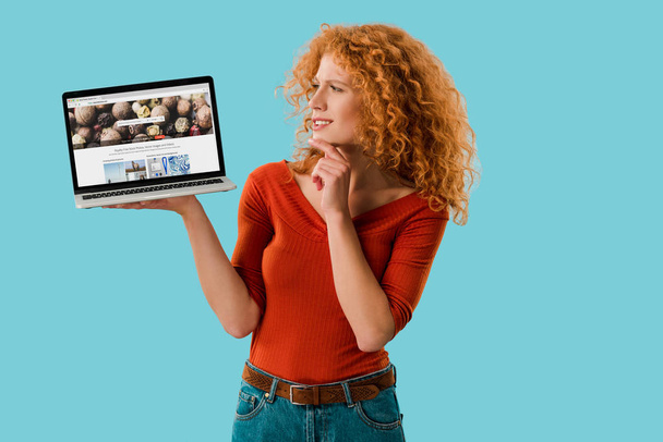 KYIV, UKRAINE - JULY 16, 2019: pensive redhead woman holding laptop with depositphotos website, isolated on blue  - Fotó, kép