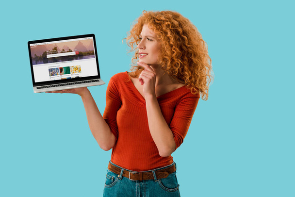 KYIV, UKRAINE - JULY 16, 2019: pensive redhead woman holding laptop with shutterstock website, isolated on blue  - Fotografie, Obrázek