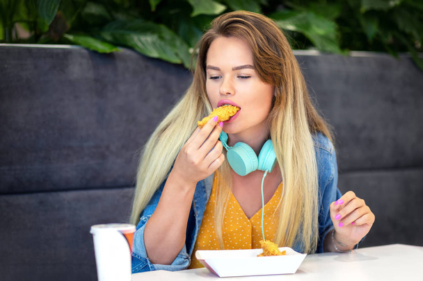 junge lächelnde Trendfrau isst knusprige Streifen beim Fast Food - fröhliches modisches Hipstermädchen in lässiger Kleidung und Headsets mit gebratenem Huhn und ungesundem Essen im Restaurant - Foto, Bild