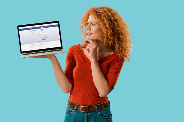 KYIV, UKRAINE - JULY 16, 2019: pensive redhead woman holding laptop facebook with website, isolated on blue  - Fotó, kép