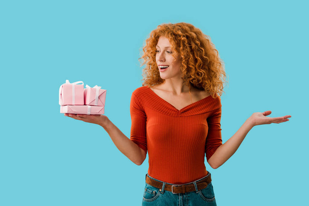 attractive redhead woman holding pink gifts isolated on blue - Photo, Image