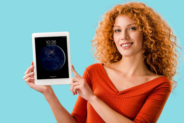 KYIV, UKRAINE - JULY 16, 2019: smiling redhead woman holding digital tablet with ipad, isolated on blue  - 写真・画像
