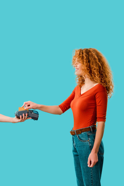 encaracolado ruiva mulher pagando com cartão de crédito no terminal, isolado em azul
 - Foto, Imagem
