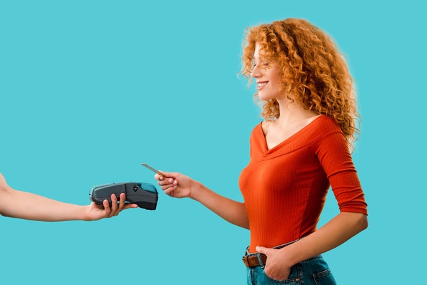 lachende redhead vrouw betalen met creditcard op de Terminal, geïsoleerd op blauw - Foto, afbeelding