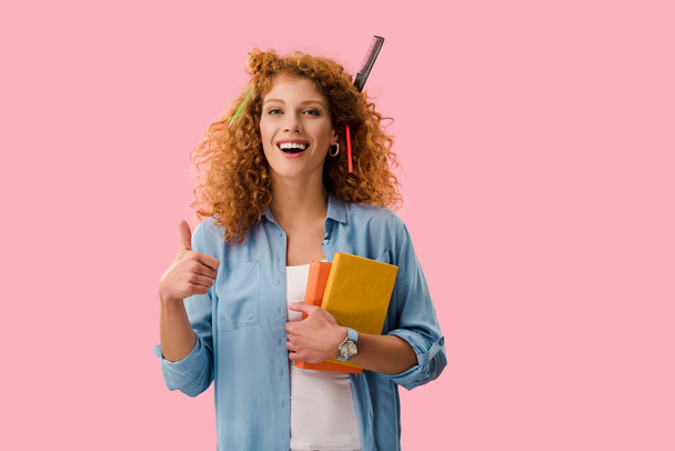 redhead student with pencils in hair holding books and showing thumb up isolated on pink - Photo, Image