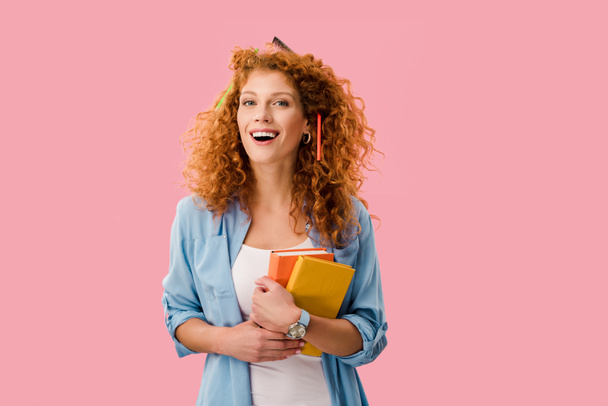 happy redhead student with pencils in hair holding books isolated on pink - Photo, Image