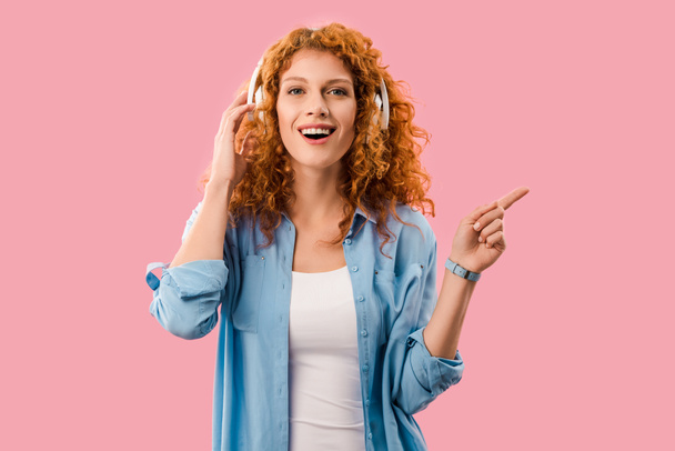curly redhead girl gesturing and listening music in headphones, Isolated On pink - Фото, изображение