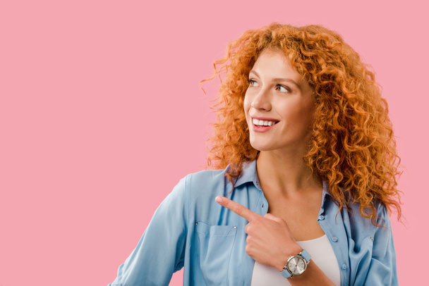 attraktive, fröhliche rothaarige Frau zeigt auf etwas Isoliertes auf rosa - Foto, Bild