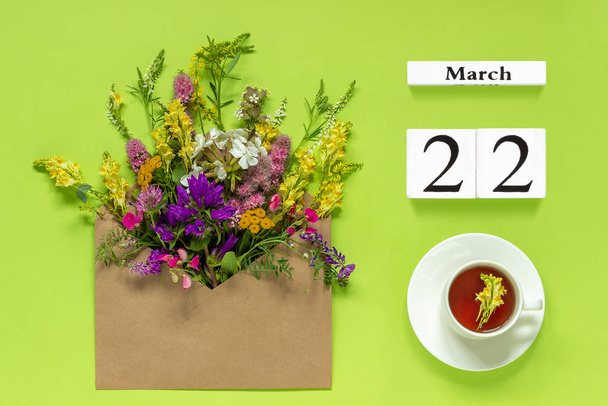 Wooden cubes calendar March 22. Cup of herbs tea, craft envelope with multi colored flowers on green background - Fotó, kép