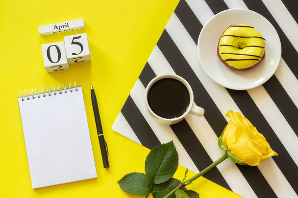 Wooden cubes calendar April 5th. Cup of coffee, yellow donut and rose on black and white napkin, empty open notepad for text on yellow background - Photo, Image