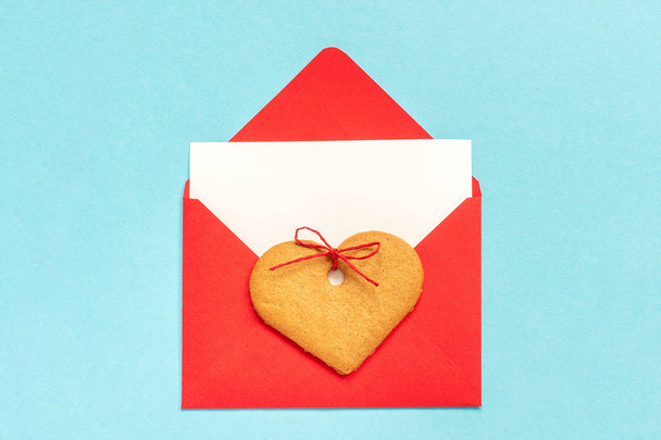 Red envelope with blank white card for text and heart shaped ginger cookie on blue background - Zdjęcie, obraz