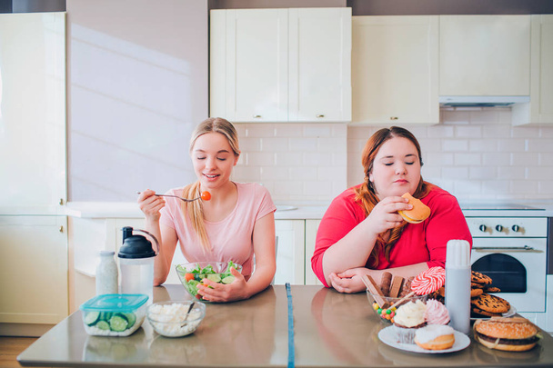 Young slim and overweight women sit at table in kitchen and eat food. Healthy and unhealthy side. Loosing weight or not. Body positive. - Foto, Imagen