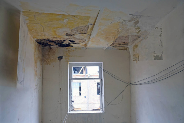 修理が必要な古い建物。屋根の損傷や漏れ後の古い住居。住宅の入り口に窓が付いているギザギザの天井と白い壁. - 写真・画像