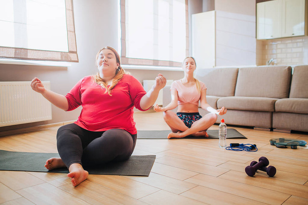 Calm peaceful young slim and plus size models sit in lotus pose and meditating. Together in living room. Workout for two. Dumbbells on floor. - 写真・画像