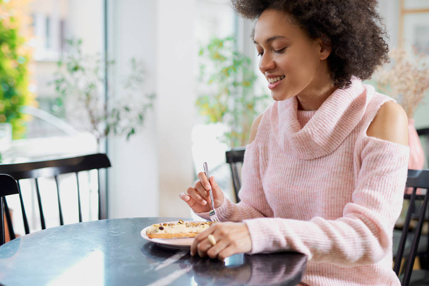 菓子に座ってガトーを食べるピンクのタートルネックセーターで魅力的なポジティブ混合レース女性の肖像画. - 写真・画像
