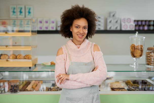 Beautiful smiling mixed race female owner of pastry shop dressed in apron standing with arms crossed in shop. - Φωτογραφία, εικόνα