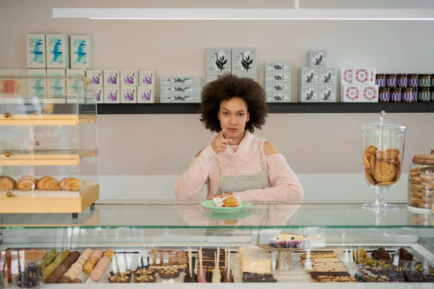 Atraktivní smíšený závod v cukrárně, ženský pracovník stojící vedle pultu a ukazující na kameru. - Fotografie, Obrázek