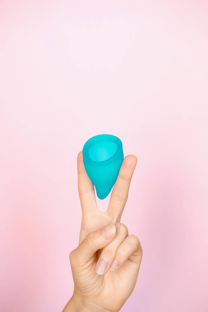 Mulher segurando em dois dedos copo menstrual azul no fundo rosa. Higiene feminina produto alternativo em vez de tampão durante o período. Menstruação, dias críticos, períodos femininos. Resíduos zero
 - Foto, Imagem
