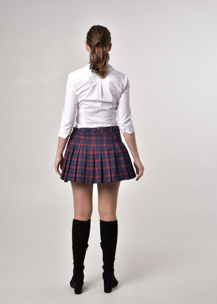 full length portrait of a brunette girl wearing a red leather jacket and plaid skirt, standing pose with back to the camera on a cream studio background. - Photo, Image