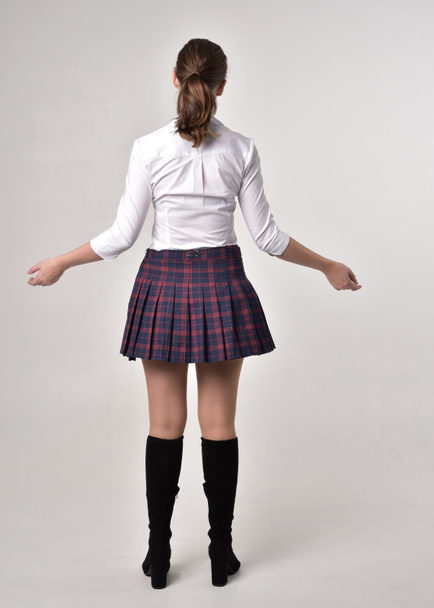 full length portrait of a brunette girl wearing a red leather jacket and plaid skirt, standing pose with back to the camera on a cream studio background. - Photo, Image