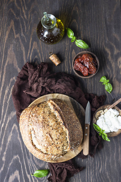 Round loaf of freshly backed sourdough bread with ricotta cheese, dried tomatoes, basil and olive oil. Dark wooden background, rustic style.  - Foto, Imagen