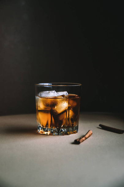 Whiskey with ice cubes and cinnamon stick on dark background. Autumn alcohol concept - Photo, Image