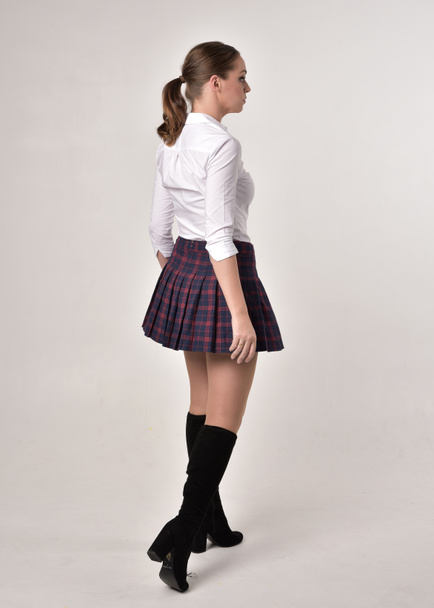 full length portrait of a brunette girl wearing a red leather jacket and plaid skirt, standing pose with back to the camera on a cream studio background. - Foto, Imagen