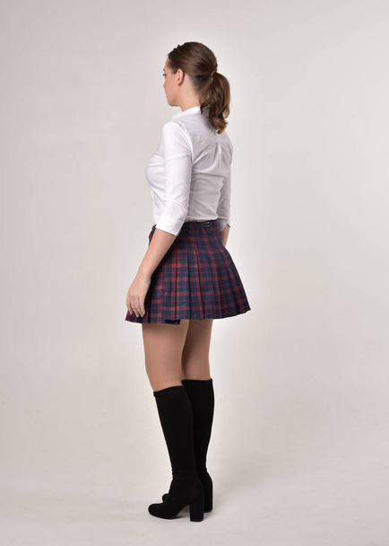 full length portrait of a brunette girl wearing a red leather jacket and plaid skirt, standing pose with back to the camera on a cream studio background. - Zdjęcie, obraz