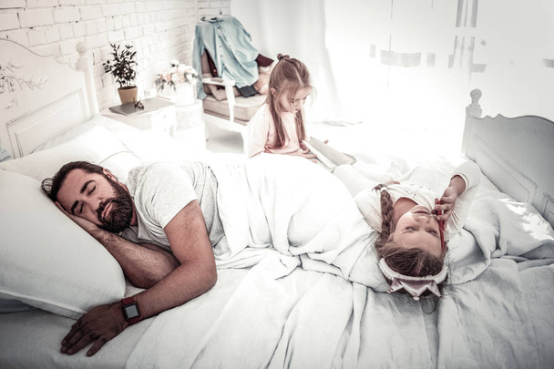 Ojciec śpi, podczas gdy jego dzieci bawią się na łóżku - Zdjęcie, obraz