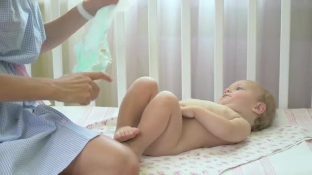 Young woman wears a diaper on the baby. Young woman sit near the baby on the cot. Little girl stands on feet in a baby cot. Mom fixes diaper on the little girl. - Video