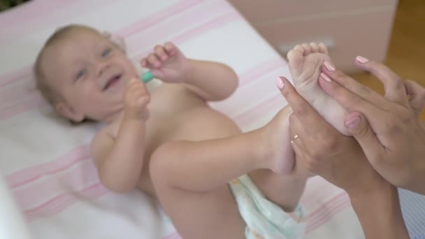 Mam maakt massage van benen voor haar baby. Een vrouw houdt een baby voet vast en wrijft het in. Klein meisje liggend op het bed. Jonge vrouw zit naast de baby op het bed. Mam speelt met de baby in een babybedje. - Video