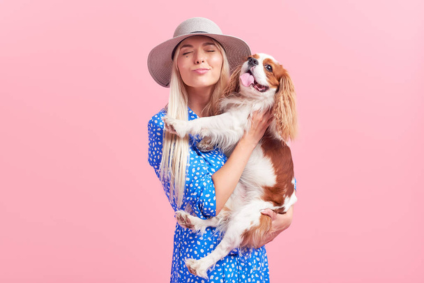 Portrait of smiling young blond woman in summer hat embracing king Charles spaniel dog. owner and pet relations concept. Veterinary health. Isolated front view on pink background. - Foto, immagini