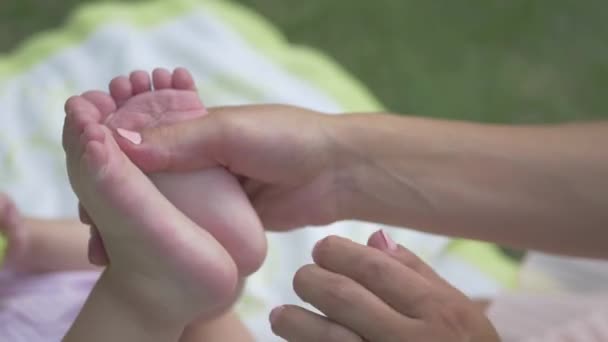 Mama macht dem Baby eine Fußmassage im Garten. Frau reibt Babyfüße. Mama massiert ihrem Baby die Beine. Kleines Mädchen liegt auf einer Decke im Garten. - Filmmaterial, Video