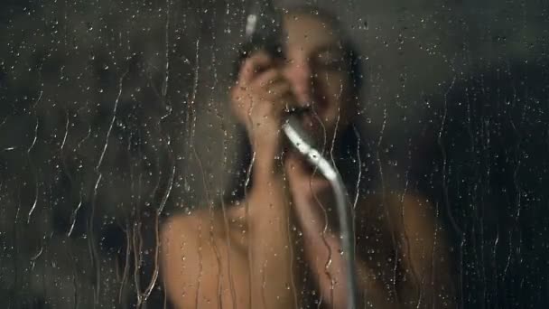 Gülümseyen genç kadın eğleniyor ve bathromm camına su döküyor. Cam ve su damlaları odaklanın. Slowmotion. - Video, Çekim