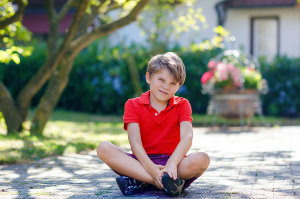 Portrait of little cool kid boy sitting on ground on sunny day in domestic backyard. Happy healthy child having fun on warm sunny day. Family, nature, love and active leisure. Kid in school uniform - Photo, Image