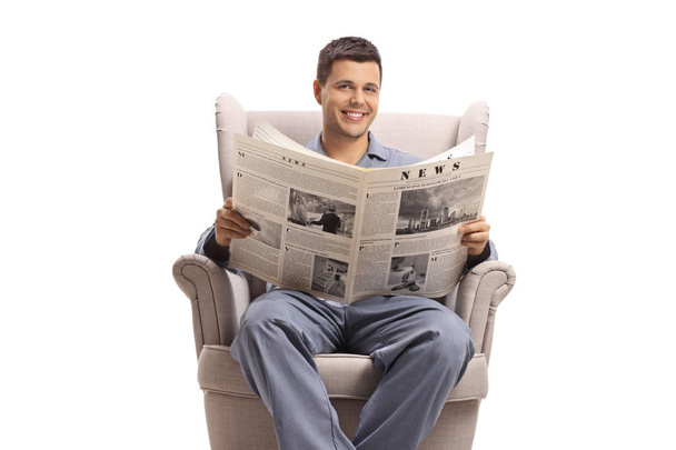 Young man in pajamas seated in an armchair with a newspaper - Фото, зображення