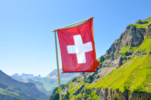 Σημαία της Ελβετίας με καλοκαιρινό ορεινό τοπίο στο παρασκήνιο. Εθνικό σύμβολο. Ελβετικές Άλπεις. Λευκός Σταυρός σε κόκκινο πεδίο. Εθνική ιδέα. Ελβετία καλοκαίρι - Φωτογραφία, εικόνα
