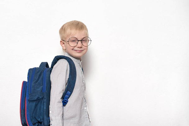 眼鏡をかけた金髪の少年の肖像画と、白い背景に学校のリュックサックを持つ。学校のコンセプト - 写真・画像