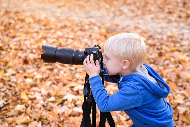 Portret van een kleine blonde jongen met een grote DSLR camera op een statief. Foto sessie in het herfst Park - Foto, afbeelding
