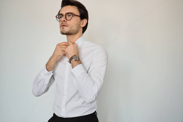 Joven atractivo morena seria hombre en camisa blanca y gafas con reloj cuidadosamente mirando a un lado sobre fondo blanco
 - Foto, Imagen