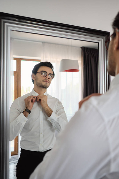 Jonge knappe brunette man in wit shirt, zwarte broek en brillen zorgvuldig op zoek in spiegel bij modern huis - Foto, afbeelding