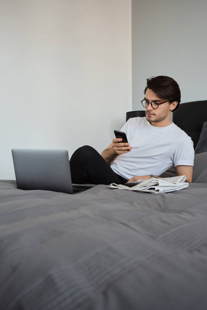 Молодий красивий серйозний брюнетка людина в білій футболці і окулярах задумливо використовує мобільний телефон, лежачи на великому ліжку з ноутбуком і газетами в сучасному будинку
 - Фото, зображення