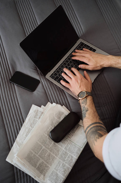 Close up photo of man hands with tattoo working on laptop with cellphone, newspapers and case for eyeglasses near on bed at home - Fotografie, Obrázek