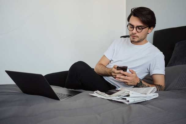 Jovem homem morena bonito em camiseta branca e óculos cuidadosamente usando celular deitado na cama com laptop e jornais na casa moderna
 - Foto, Imagem