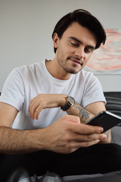 Fiatal jóképű mosolygó barna férfi fehér póló álmodosan használ cellphone ül az ágyon laptop a modern otthon - Fotó, kép